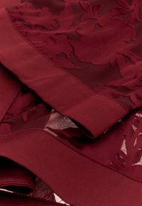 Bordeaux stof met een gelaagd ontwerp dat bloemenpatronen bevat, een gladde textuur en contrasterende accenten. De randen zijn netjes gestikt.