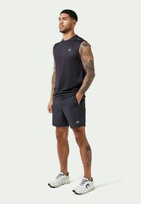 Athletic man with tattoos stands wearing black sleeveless shirt, black shorts, and white running shoes on plain light background.