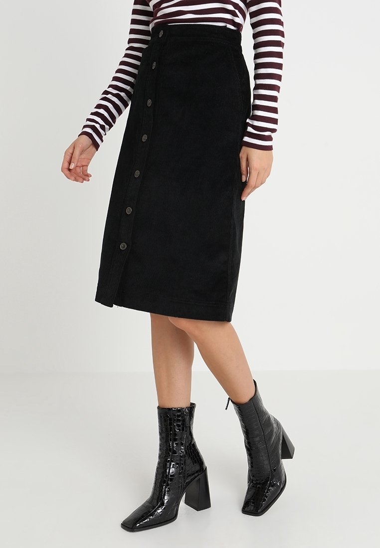 Femme portant une jupe midi en velours côtelé noire boutonnée, un haut à manches longues rayé, et des bottines noires à talons texturées.