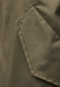 Κοντινή λήψη υφάσματος ελιάς με ραμμένη, γωνιώδη τσέπη που διαθέτει διακριτό υφή με μοτίβο πλέγματος.