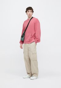 Ροζ φούτ�ερ με μπροστινή τσέπη, συνδυασμένο με μπεζ παντελόνια cargo και λευκά αθλητικά παπούτσια. Στολισμένο με πράσινη τσάντα χιαστί.