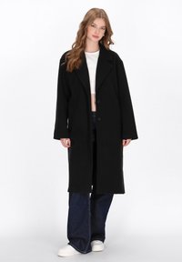 Abrigo largo negro con cuello de muesca, cierre de un solo botón y dos bolsillos laterales; llevado sobre un top corto blanco y combinado con jeans acampanados.