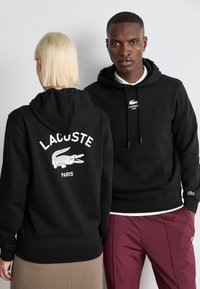 Black hoodie featuring a white Lacoste logo and crocodile graphic on the back. Soft fabric, front pocket, and drawstring hood.