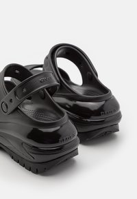Zuecos Crocs negros y brillantes con suelas texturizadas y correas ajustables en el talón, mostrados desde un ángulo lateral trasero sobre un fondo blanco.