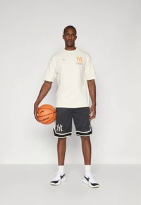 Világosbézs póló narancssárga NY logóval, fekete hálós rövidnadrággal, fehér szegéllyel és NY emblémával. Kosárlabdát tart a kezében.