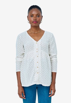 Frau trägt weiße, geknöpfte V-Ausschnitt-Strickjacke mit Zickzackmuster und blaue Jeans, steht vor hellem einfarbigem Hintergrund.