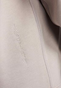 Tissu sportif beige clair avec une texture lisse, featuring le logo en relief "THERM-A-FIT", des détails de couture contrastants et une coupe ajustée.
