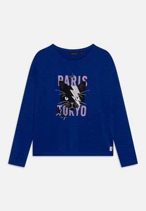 Blauw shirt met lange mouwen met een zwart kattengezicht dat een zilveren bliksemschicht draagt, roze "PARIS" boven en "TOKYO" onder met zwarte steraccenten.