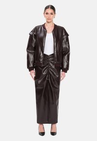 Giacca bomber marrone in ecopelle con colletto a coste, abbinata a una gonna lunga arricciata coordinata. Tacchi neri completano il look.