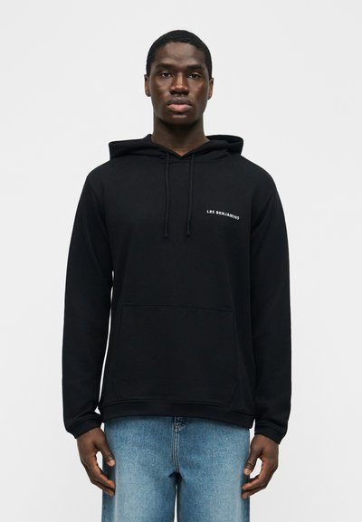 Άνδρας με μαύρο φούτερ Les Benjamins και μπλε τζιν, στέκεται μπροστά σε απλό φωτεινό φόντο.