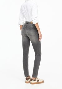 Mujer vistiendo jeans ajustados grises, camisa blanca con mangas arremangadas y zapatillas beige con detalles marrones, de pie contra un fondo blanco.