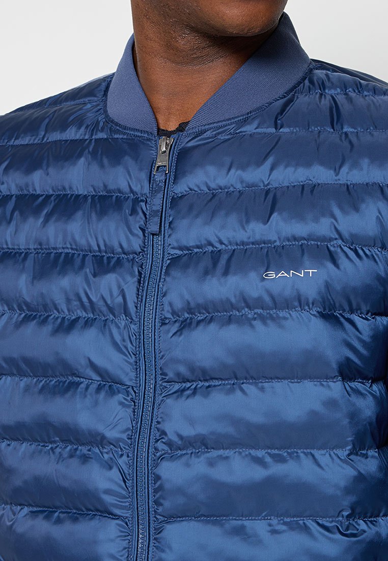 Niebieska, pikowana kurtka puchowa z zamkiem z przodu, żebrowanym kołnierzem i przeszyciami. Posiada małe logo na piersi.