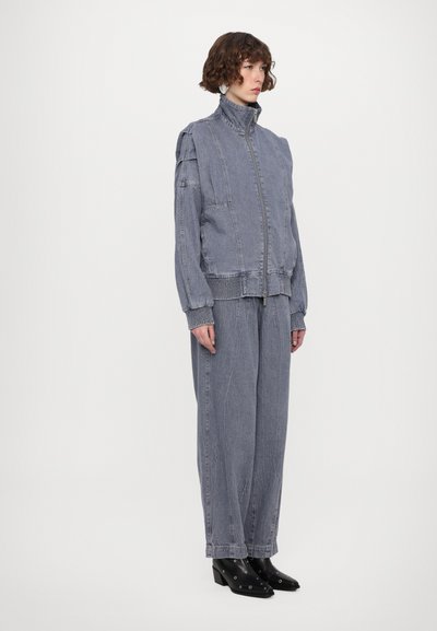 Jachetă din denim de culoare albastru decolorat, cu fermoar centrat, guler înalt și manșete ribbate. Asortată cu pantaloni din denim largi și cizme până la glezne.