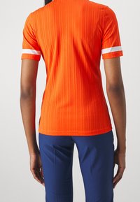 Ribbad orange kortärmad topp med vita accentremsor vid ärmsluten, med en figursydd design och slät textur, kombinerad med marinblå byxor.