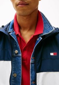 Chaqueta bloqueada en azul marino y blanco con detalles de mezclilla y herrajes plateados, superpuesta sobre una camiseta polo roja con un acabado texturizado.