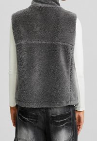 Szara polarowa kamizelka z wysokim kołnierzem, miękką teksturą i przeszyciem na plecach. Nosi się ją na lekki top z długim rękawem w połączeniu z dżinsami.