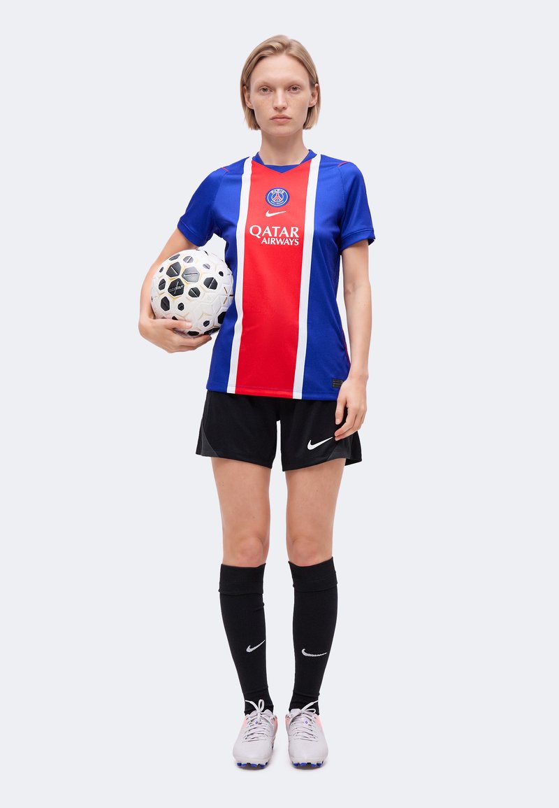 Jeune joueuse de football en maillot du Paris Saint-Germain tenant un ballon de football blanc et noir, portant un short, des chaussettes et des chaussures noires.