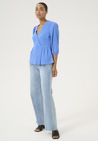Top avvolgente azzurro chiaro, con texture, maniche a sbuffo e orlo peplum; abbinato a jeans in denim a gamba larga e décolleté nere aperte.