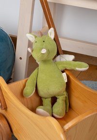 Green plush dragon toy with a soft texture, white accents, small wings, and a smiling face, seated in a wooden cart.