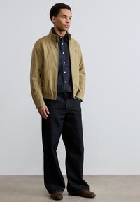 Νεαρός άνδρας όρθιος με τα χέρια στις τσέπες, φοράει ταμπά ζακέτα με φερμουάρ, σκοτεινό πουκάμισο με κουμπιά, φαρδιά μαύρα τζιν και καφέ παπούτσια μπροστά σε απλό φόντο.