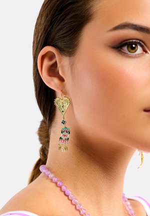 Gold heart-shaped earring with intricate cutouts and multi-colored beads, featuring a dangling design. Pink beaded necklace visible.