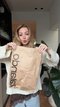 Brūna papīra maisiņš ar melniem burtiem "ob", ko tur persona, valkājot krēma krāsas džemperi. Fonā redzama zaļa augs un plaukti ar produktiem.