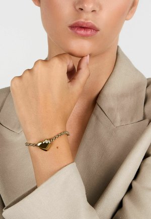 Bracciale dorato con un ciondolo a forma di cuore e chiusura decorativa, caratterizzato da un design a catena testurizzata. Indossato su un polso contro uno sfondo di tessuto chiaro.