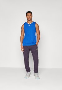 Zila bezsleeve sporta blūze ar baltajiem akcentiem, apvienota ar tumši violetām biksēm un pelēkiem skriešanas apaviem. Vienkāršs, funkcionāls sporta apģērba dizains.