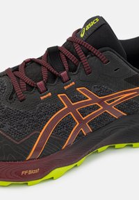 Svart atletisk sko med teksturert stoffoverdel. Har oransje og mørkerøde detaljer, reflekterende elementer og gult logo. Grønn gummisåle.