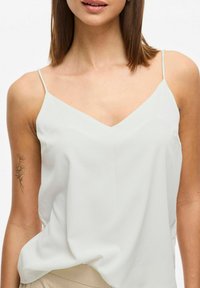 Camisole alb deschis cu decolteu în V și bretele subțiri, având o textură netedă și o croială lejeră, asociată cu material bej dedesubt.