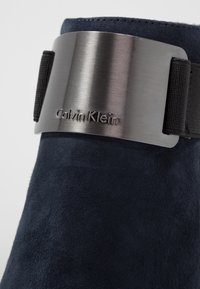 Bottine en daim bleu marine avec une bande argentée métallique embossée du logo "Calvin Klein" et une sangle élastique noire pour un ajustement sûr.