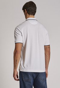 Polo shirt bianco a maniche corte con colletto e polsini in contrasto blu navy. Texture liscia, vestibilità leggermente ampia e dettaglio del marchio nella parte posteriore del collo.