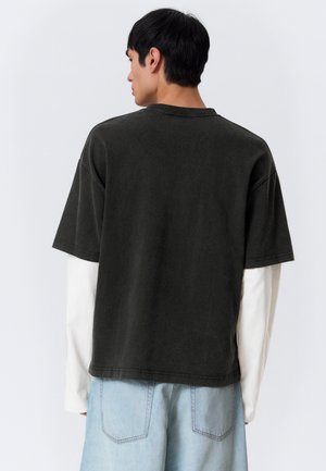 Homme portant une chemise noire à manches courtes par-dessus une chemise blanche à manches longues et un jean bleu clair, vu de dos sur un fond uni.