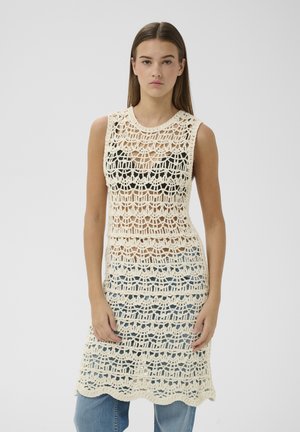 Joven mujer con vestido de crochet crema sin mangas sobre top negro y vaqueros azules, de pie frente a un fondo claro y liso.