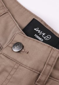 Close-up van de tailleband van beige broek met een metalen knoop met een "T" en een zwart label met "FORMER" en daarboven een handtekening.