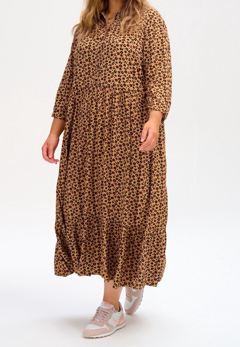 Langes, braunes Kleid mit Leopardenmuster, Stufendesign und dreiviertel Ärmeln. Kombiniert mit hellrosa Sneakern. Glatte Stoffstruktur.