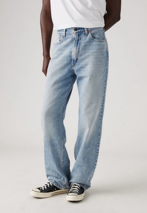 Personne portant un jean bleu clair ample, un t-shirt blanc et des baskets noires basses, debout devant un fond uni.