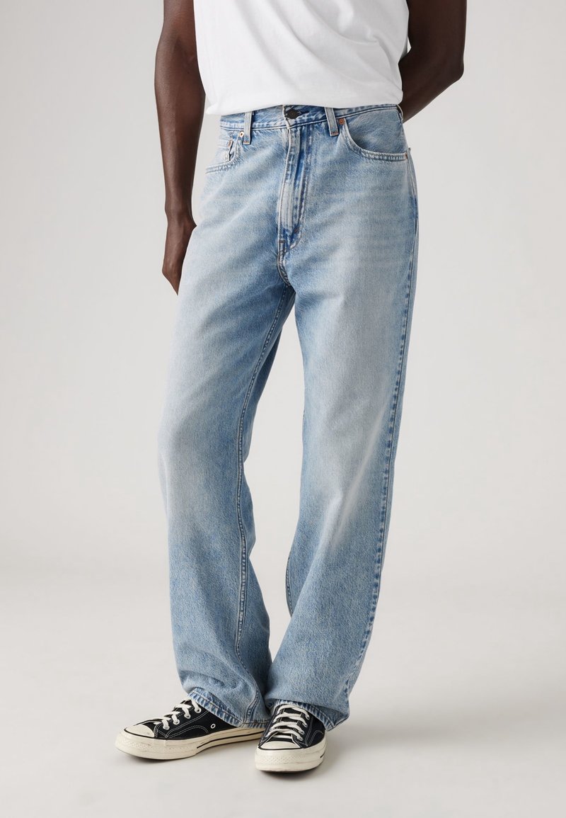 Personne portant un jean bleu clair ample, un t-shirt blanc et des baskets noires basses, debout devant un fond uni.