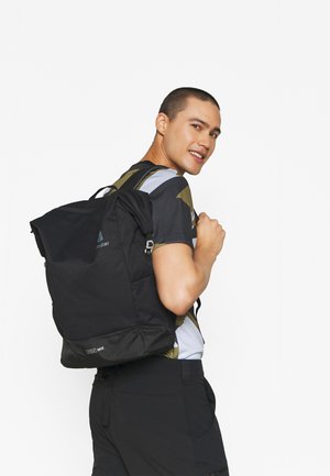 Junger Mann, der über die Schulter lächelt, eine große schwarze Tasche trägt, ein gemustertes kurzärmliges Hemd und schwarze Hosen trägt, vor einem weißen Hintergrund.