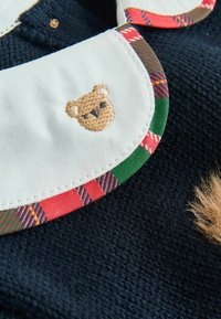 Marine textiel met een witte kraag, voorzien van een geborduurd beer en een kleurrijke ruitrand in rode, groene en zwarte accenten.