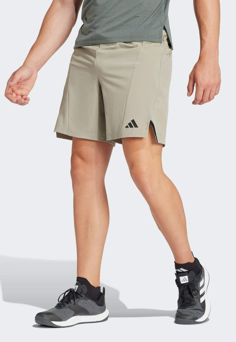 Beige Shorts mit glatter Textur, ausgestattet mit seitlichen Schlitzen und einem schwarzen Adidas-Logo. Kombiniert mit schwarzen und grauen Sportschuhen.