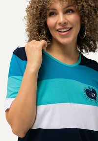 Farverig stribet T-shirt med blå og hvide striber, rund halsudskæring og et hjerte design med et øje, lavet af blødt stof.