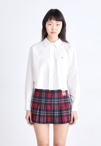 Camisa blanca tipo cropped con cuello, combinada con una falda plisada de cuadros en rojo, azul y verde que presenta una etiqueta en la cinturilla.