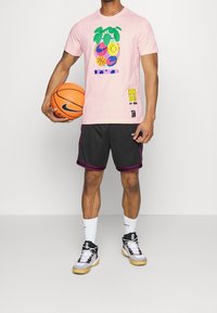 Camiseta gráfica rosa con patrones de frutas coloridas y caritas sonrientes, combinada con pantalones cortos grises. Zapatillas blancas y un balón de baloncesto completan el look.