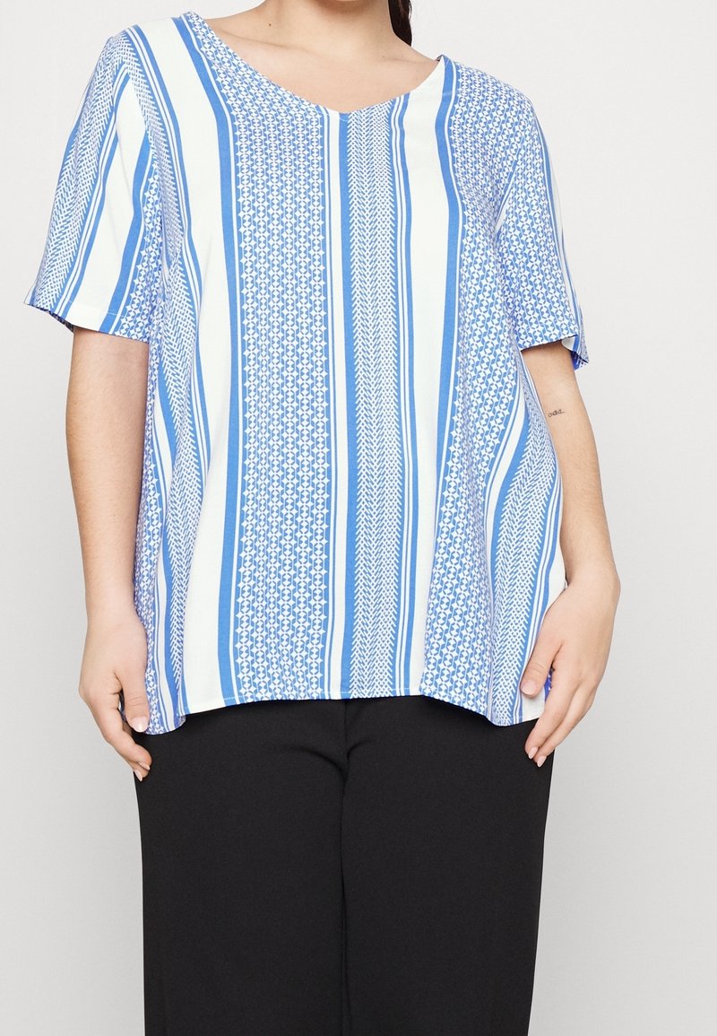 Nainen, jolla on päällään löysä sinivalkoraidallinen lyhythihainen paita ja mustat housut, seisoo kädet rentona sivuilla.