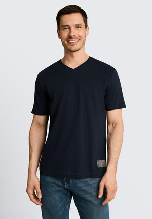 Lachende man die een effen zwart V-hals T-shirt en blauwe jeans draagt, staand tegen een lichtgrijze achtergrond.