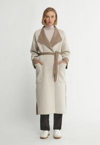 Lång, ljusgrå kappa med brun krage och bälte, tillverkad av mjukt tyg; har stora fickor och sidoklyftor. Buren över mörka byxor och vita skor.