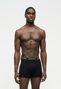 Μυώδης άνδρας όρθιος, φοράει μαύρα ανδρικά εσώρουχα Björn Borg, κοιτάζοντας μπροστά σε απλό φωτεινό φόντο.