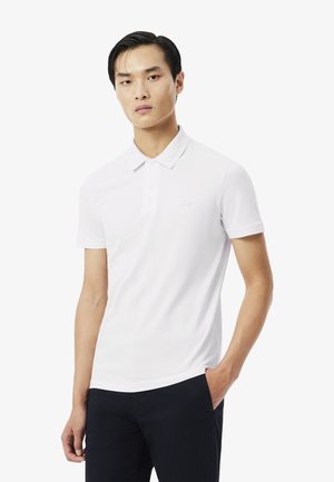 Giovane uomo che indossa una polo bianca a maniche corte e pantaloni scuri, in piedi con una mano in tasca contro uno sfondo semplice.