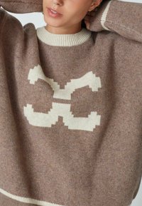 Smeđi pleteni pulover s kremastim geometrijskim uzorkom, rebrasti izrez i manšete, oversized kroj i mekana, teksturirana tkanina.
